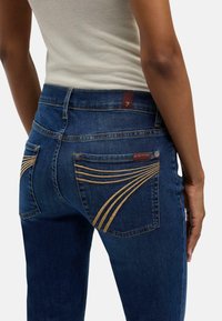 Blå jeans i denim med kontrasterande gyllene sömmar på bakfickorna. Har en brun etikett på midjebandet och en figursydd design.