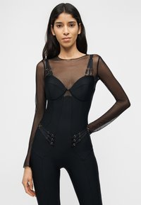 Γυναίκα που φοράει εφαρμοστό μαύρο ολόσωμο φόρεμα με διάφανα μακριά μανίκια και διακοσμητικές λουρίδες στο στήθος και τους γοφούς, ποζάροντας με το ένα χέρι στη μέση.