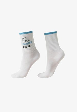 Calcetines blancos de tripulación con ribete azul, uno con el texto: "COME. DUERME. PILATES. REPITE." en letras negras y azules.