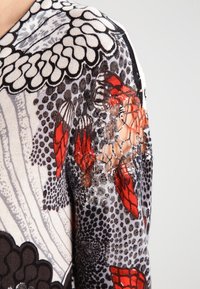 Nära bild av ett långärmat plagg med svarta, vita och röda fjärilar och abstrakta mönster samt en spetsdetalj på överarmen.
