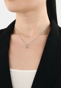 Silver kedja halsband med en liten grön ädelsten hängande på en person klädd i en svart blazer och vit topp.