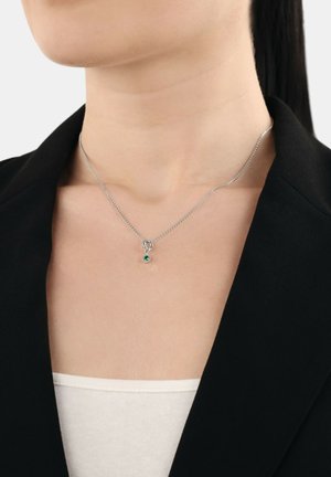 Collana in argento con un piccolo pendente in pietra verde indossata da una persona vestita con un blazer nero e una maglietta bianca.