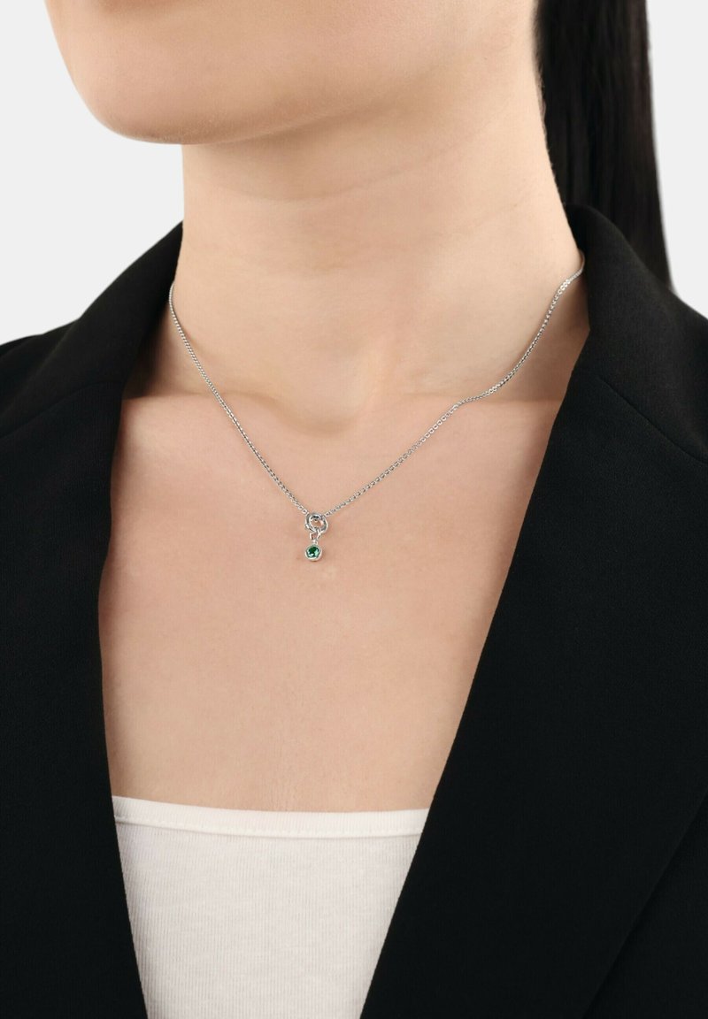 Silver kedja halsband med en liten grön ädelsten hängande på en person klädd i en svart blazer och vit topp.