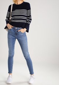 Mujer vistiendo un suéter de rayas azul marino y blanco, jeans ajustados de color azul claro, zapatillas blancas y un bolso de hombro beige, con las manos en los bolsillos.