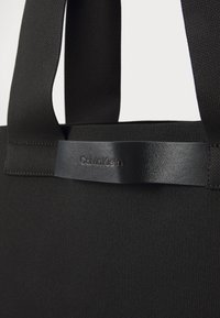 Must must ülekanne, millel on kangast keha ja sile must nahkdetail. Omab pehmet tekstuuri, vastupidavaid rihmu ja reljeefset "Calvin Klein" logo.