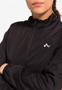 Czarna lekka kurtka z wysokim kołnierzem, przednim zamkiem błyskawicznym i elastycznymi mankietami. Posiada małe białe logo na lewej stronie klatki piersiowej.