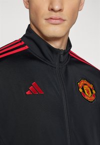 Sort zip-up jakke med røde accenter, med tre striber på skuldrene, broderet Manchester United-logo og Adidas-logo på brystet.