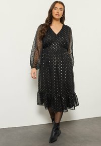 Musta mekko, jossa on läpinäkyvät puffihihat, kultaiset pistekuviot, V-hihapääntie, röyhelöhelma ja istuva vyötärö. Yhdistetty mustiin nilkkabootseihin.