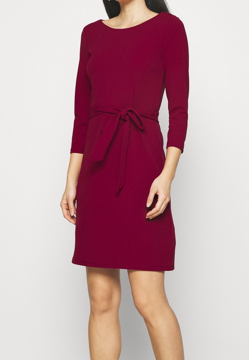 Une femme porte une robe bordeaux longueur genou avec des manches trois-quarts et une ceinture nouée à la taille, sur un fond blanc uni.