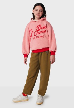 Rosa Kapuzenpullover mit rotem Text, lockere Passform, weißes Innenfutter, rote Bündchen. Olivgrüne Hose, weicher Stoff, kombiniert mit weißen Sneakers.