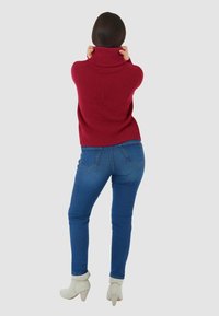 Maglione a collo alto rosso in maglia abbinato a jeans di denim blu. L'outfit presenta una vestibilità rilassata e una texture a coste, completato da stivaletti beige alla caviglia.
