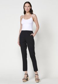 Pantalon noir ajusté avec une coupe slim, doté de poches latérales et d'une taille plate, associé à un débardeur blanc ajusté et des chaussures compensées.