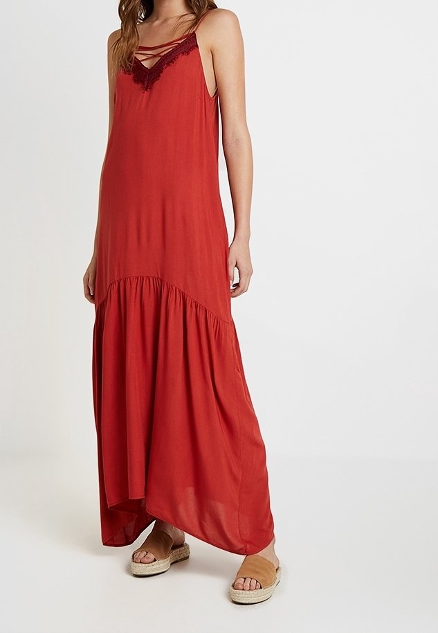 Sukienka maxi w kolorze rdzawym, z koronkowymi detalami i falbanami na dole. Gładka tekstura z regulowanymi ramiączkami, w połączeniu z neutralnymi sandałami.