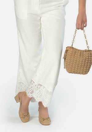 Mujer con pantalones blancos con ribete de encaje, zapatos con tacón cuadrado marrón claro y hebillas doradas, sosteniendo un bolso tejido marrón claro con correa de cadena dorada.