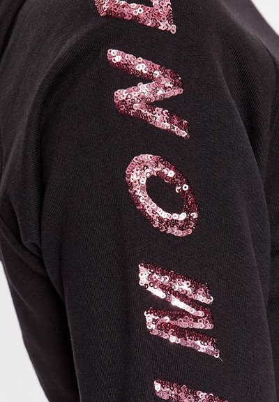 Tessuto nero con lettere di paillettes rosa verticali che formano la parola "AMON" su una manica o sulla gamba di un pantalone.
