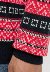 Cuff di un maglione a maglia con un motivo rosso brillante e forme geometriche, accentuato da strisce nere e bianche, realizzato in filato morbido.