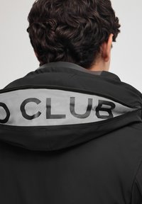 Giacca nera con cappuccio, caratterizzata da una fodera interna grigia con la scritta "CLUB" in bold nero, e una zip visibile. Tessuto liscio.