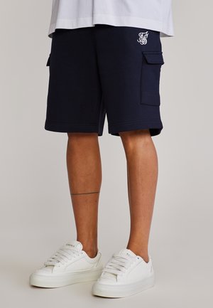 Benedenlichaam in marineblauwe cargoshorts met een logo en witte sneakers, staand tegen een effen lichte achtergrond.