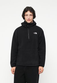 Mlađi muškarac nosi crni flis pulover s polu-zipom i kengur džepom, na kojem se nalazi bijeli logo The North Face na prsima.