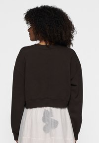 Felpa marrone corta con maniche ampie, tessuto texturizzato e scollatura rotonda, indossata su un vestito bianco trasparente con un motivo grigio.