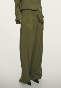 Un pantalon large taille haute vert olive en tissu texturé, avec des plis et des poches latérales, assorti d'une veste. 
