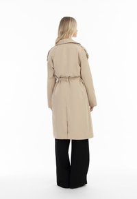 Beige trenchcoat s opaskou v pase, má zadní větrací otvor, epaulety a hladkou texturu. V kombinaci s černými kalhotami s širokými nohavicemi.