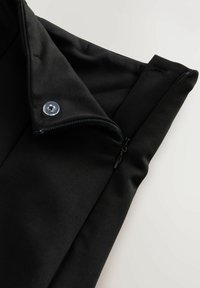 Black garment fabric with a partially opened zipper and a single translucent button on a folded flap, set against a light background.