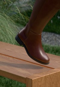 Brauner Lederstiefel bis zum Knie mit abgerundetem Zehenbereich, glatter Textur und elastischem Seitenpanel, vor einer Holzoberfläche.