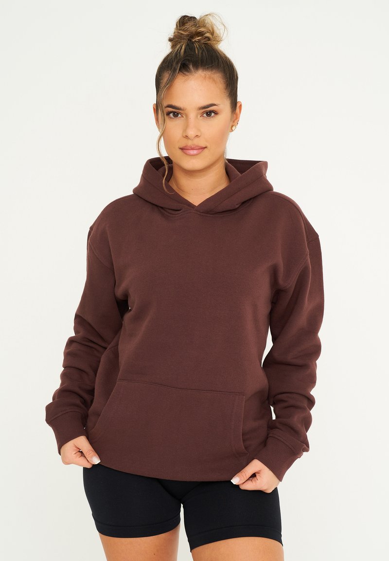 Brauner Kapuzenpullover aus weichem Stoff, mit einer Fronttasche und gerippten Bündchen. Getragen mit schwarzen Sportshorts.