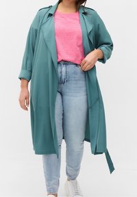 Donna vestita con un lungo cappotto turchese, una maglietta rosa, jeans azzurri chiari con strass alle caviglie e sneakers bianche, in piedi davanti a uno sfondo neutro.