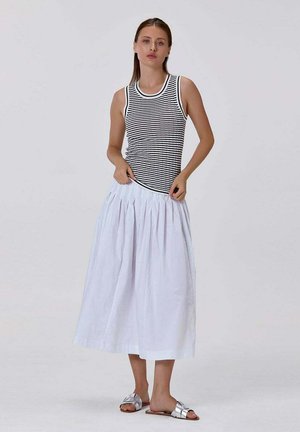 Femme debout ajustant une jupe plissée blanche, portant un haut sans manches rayé noir et blanc et des sandales argentées slide.
