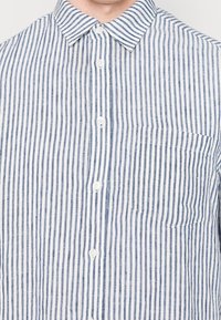 Chemise à rayures bleues et blanches en tissu léger, avec col, poche avant et surface texturée.