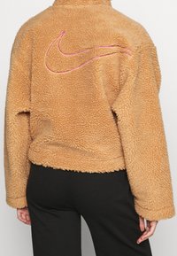 Tan fleecejacka med en fluffig textur, med en rosa broderad Nike-logotyp på baksidan, och en avslappnad passform med långa ärmar.