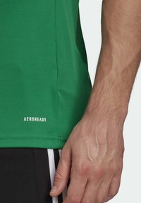 Högra handen på en person som bär en grön sporttröja med texten "AEROREADY" och svarta shorts med vita ränder.