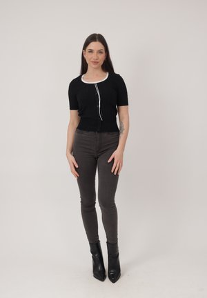 Jonge vrouw met lang donker haar, gekleed in een zwart shirt met korte mouwen, grijze skinny jeans en zwarte enkellaarzen, staand tegen een effen lichte achtergrond.