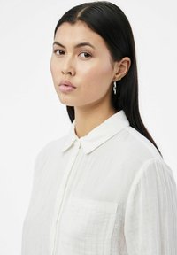 Camisa blanca con cuello, hecha de una tela texturada, que presenta un único bolsillo en el pecho y mangas largas. Modelo con cabello largo y negro y aretes de aro.