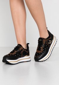 Svarta och bruna sneakers i mocka och läder, med guldfärgad dekoration, vit mellansula och dekorativa nitar på ovansidan.