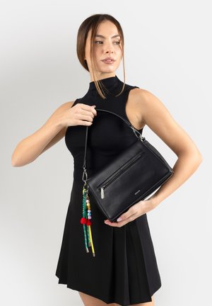 Donna in abito nero senza maniche che tiene una borsa a mano in pelle nera con un ciondolo a nappina colorato di perline sulla tracolla.