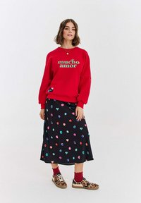 Žena v červené mikině s nápisem "mucho amor", černé sukni s barevnými puntíky, červených ponožkách a teniskách s leopardím potiskem, stojí.