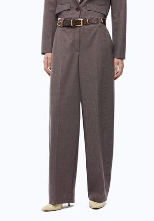 Pantalones marrones de cintura alta y pierna ancha con estampado de cuadros y cinturón oscuro, combinados con zapatos de tacón crema de punta puntiaguda, mostrados en una persona de pie.