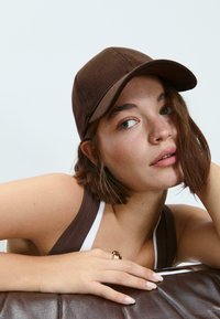 Hnedá kordurová čiapka s zakriveným okrajom, nosená nad vlasmi po plecia; má textúrovaný povrch a neformálny dizajn.