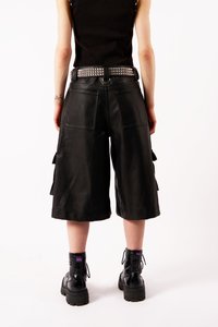 Melnās ādas kravas šorti ar brīvu piegriezumu, redzamu šuvēm un sānu kabatām. Valkāti ar studētu melnu jostu un melniem daļēji apaviem.