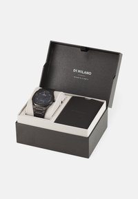 Montre noire avec un cadran rond et un bracelet métallique, présentée dans une boîte sombre avec un rembourrage blanc et un petit livret à côté.