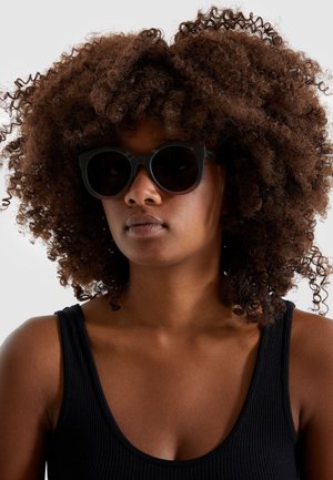 Lunettes de soleil noires avec une forme de chat arrondie, des montures épaisses et des verres sombres. Le modèle a les cheveux bruns et bouclés et porte un débardeur noir.