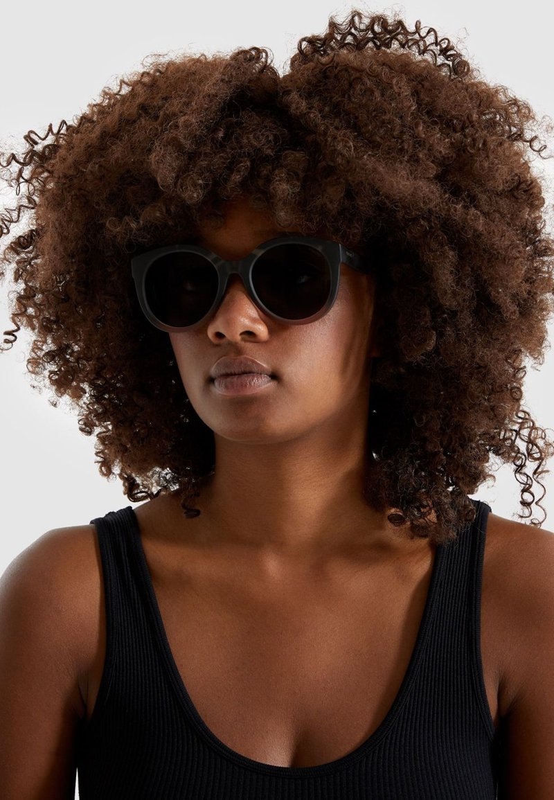 Lunettes de soleil noires avec une forme de chat arrondie, des montures épaisses et des verres sombres. Le modèle a les cheveux bruns et bouclés et porte un débardeur noir.