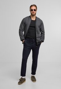 Grå zip-up jakke, sort tanktop, marineblå bukser og brune loafers med et tekstureret design. Accessories inkluderer runde solbriller.