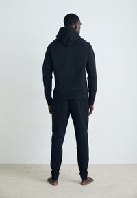 Svart hettegenser med justerbar hette, ribbede mansjetter og livkant, kombinert med løse svarte joggebukser. Myk tekstur.