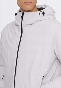 Giacca grigio chiaro realizzata in materiale morbido e impermeabile. Presenta un colletto alto, una cerniera nera, un cappuccio regolabile con dettagli rossi e tasche laterali.