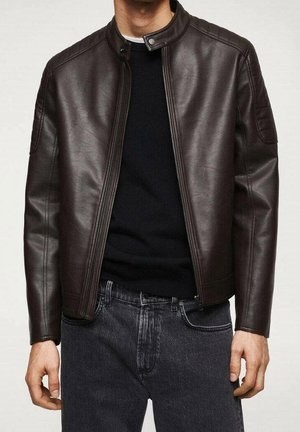 Hombre con chaqueta de cuero marrón oscuro sobre una camisa negra y jeans gris oscuro, de pie frente a un fondo claro y sencillo.