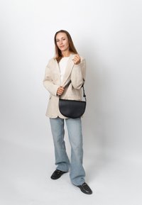 Femme debout portant un blazer beige, un haut blanc, un jean bleu clair, des chaussures noires, tenant un sac à bandoulière noir sur fond blanc uni.